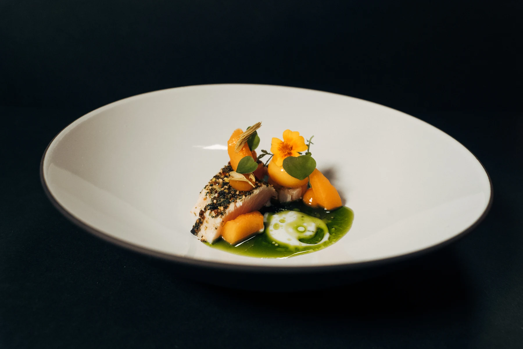 Fein angerichteter Gang mit orangefarbener Blüte und grüner Sauce vor dunklem Hintergrund